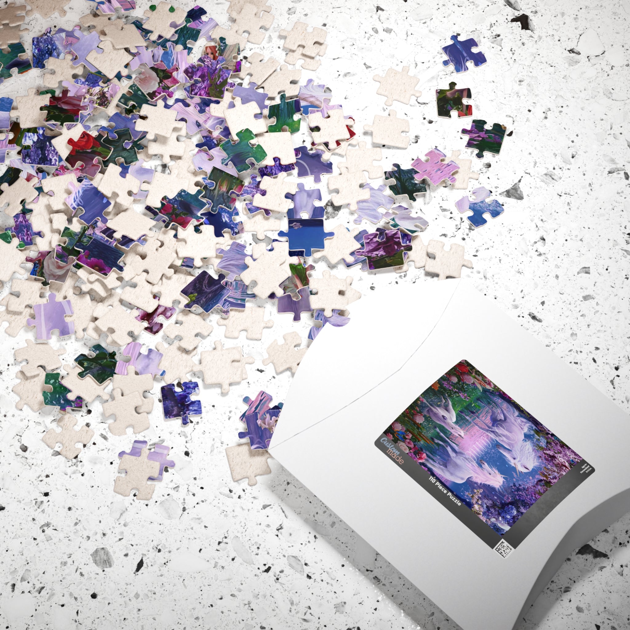 A white jigsaw puzzle box lies on a white speckled surface with many loose puzzle pieces scattered across it. The visible puzzle image shows a colorful fantasy landscape with purple, blue, pink, and green tones.