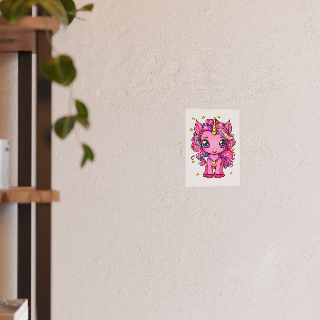 A small rectangular wall art print featuring a cute pink cartoon unicorn with a gold horn, displayed on a textured off-white wall next to a blurred wooden shelf with hanging green leaves.