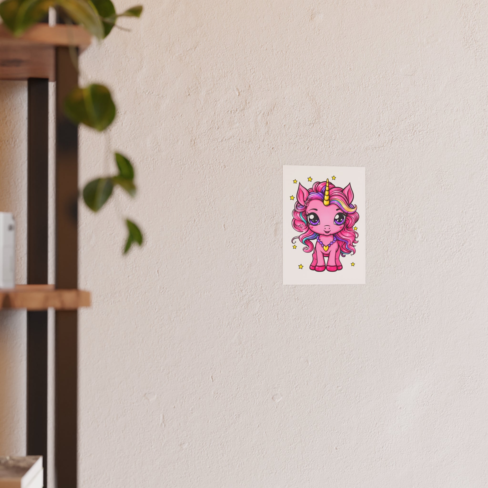 A small rectangular wall art print featuring a cute pink cartoon unicorn with a gold horn, displayed on a textured off-white wall next to a blurred wooden shelf with hanging green leaves.