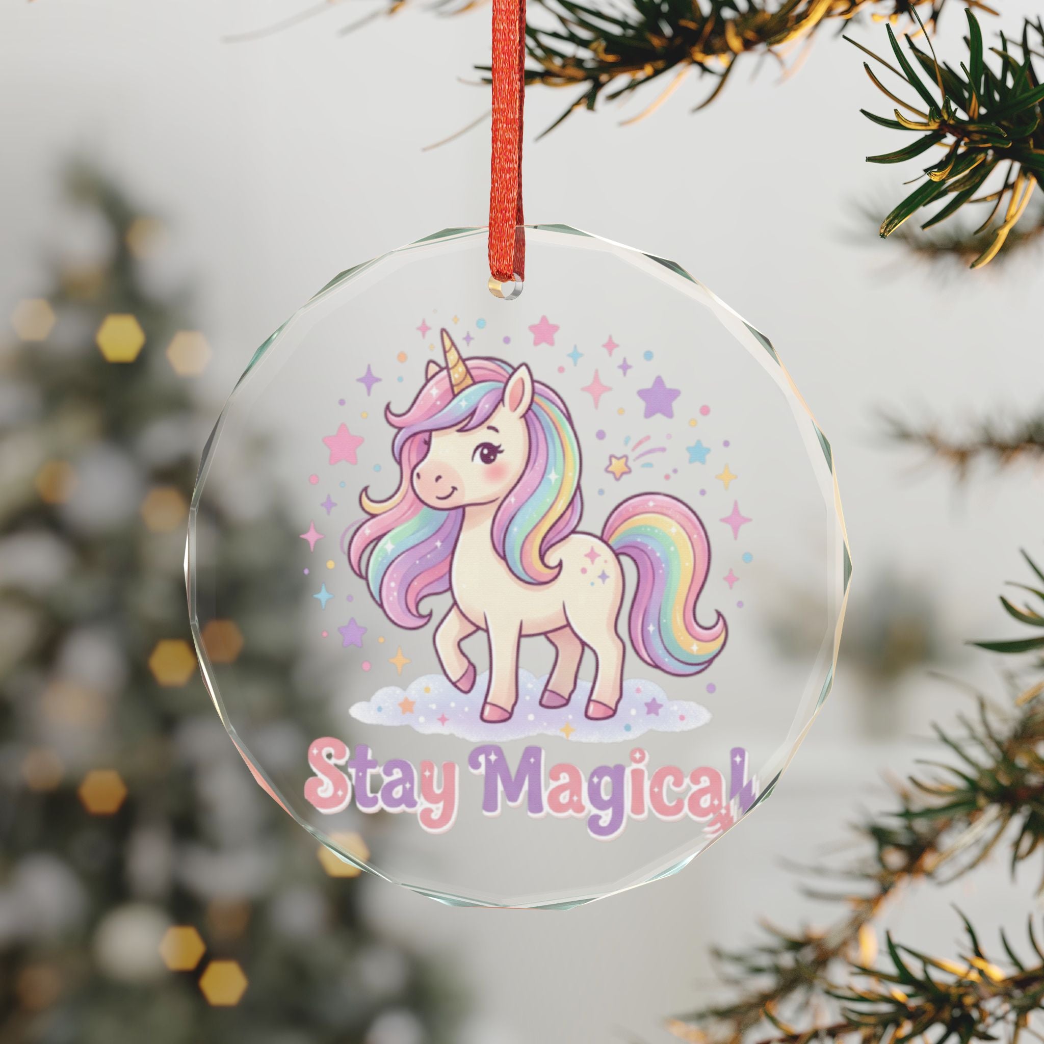 A round clear glass Christmas ornament featuring a cute white unicorn with a pastel rainbow mane and tail, surrounded by stars, with the text 'Stay Magical' at the bottom. The ornament hangs from a red ribbon and is shown hanging from a pine tree branch with a blurred holiday background.