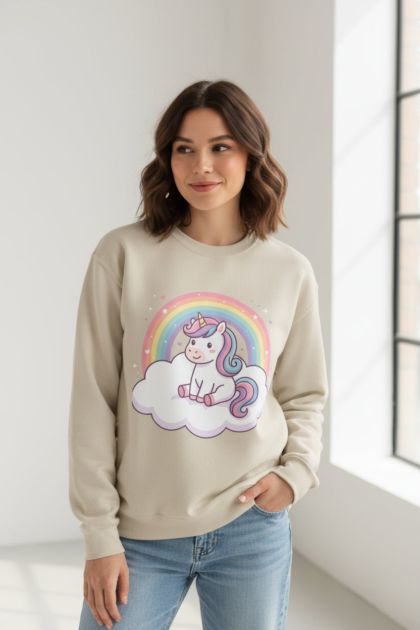 A beige crewneck sweatshirt featuring a colorful cartoon graphic of a white unicorn with a pink and blue mane sitting on a white cloud in front of a pastel rainbow.