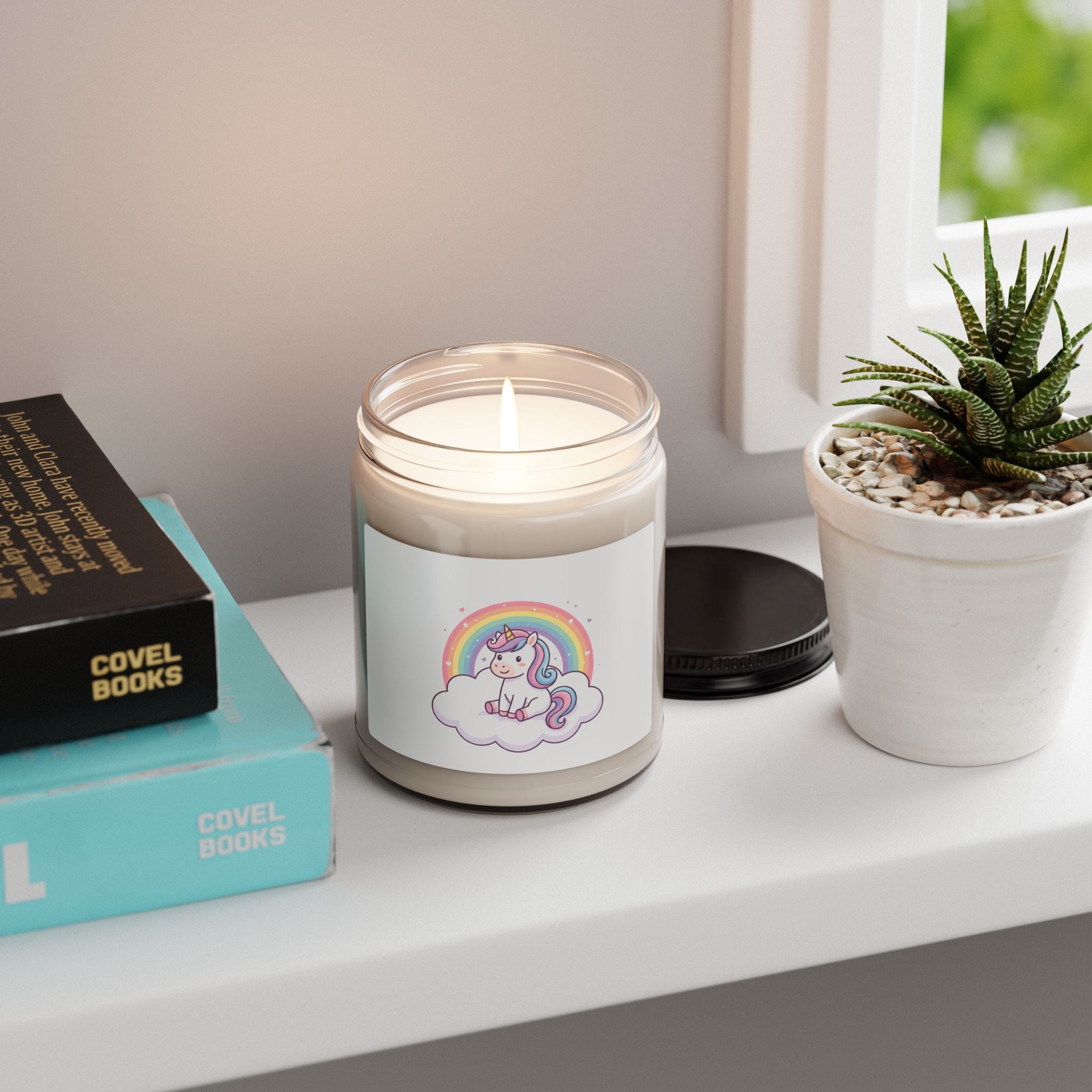 A lit candle in a clear glass jar sits on a white windowsill next to a small potted succulent and stacked books. The candle has a white label featuring a cute pastel unicorn under a rainbow.