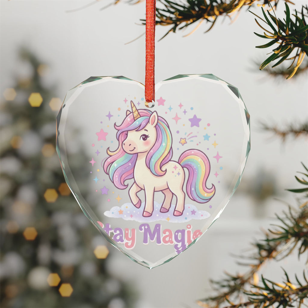 A heart-shaped clear glass Christmas ornament featuring a cute pastel unicorn with a rainbow mane and the words 'Stay Magic' at the bottom, hanging from a red ribbon on a Christmas tree branch.