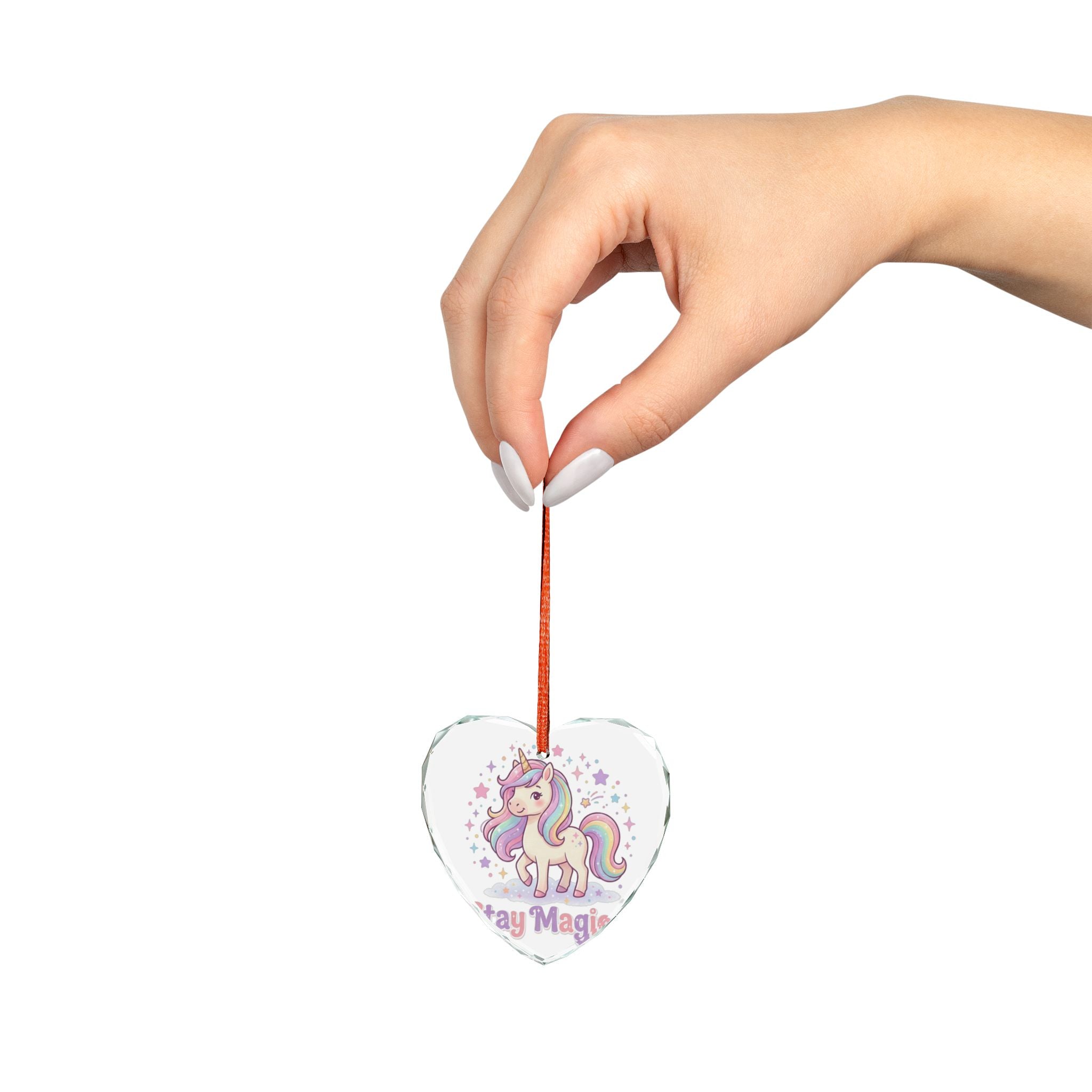 A hand with white manicured nails holds a clear heart-shaped acrylic ornament featuring a colorful unicorn illustration and the words 'Stay Magic' against a plain white background.